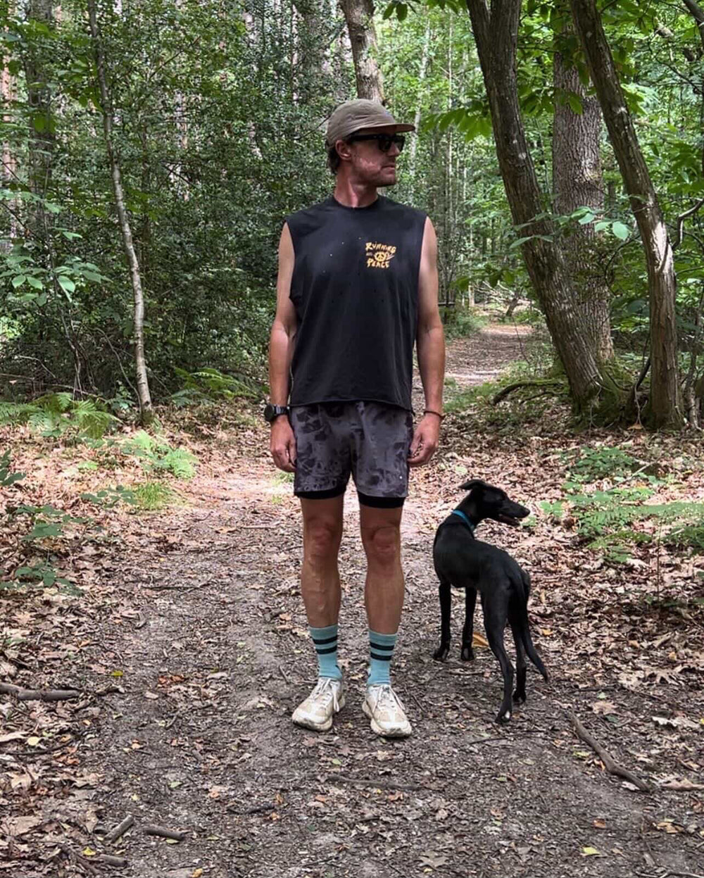 Man running on a trail in a forest wearing a black Nowhere Somewhere Running Peace tank top with yellow text.
