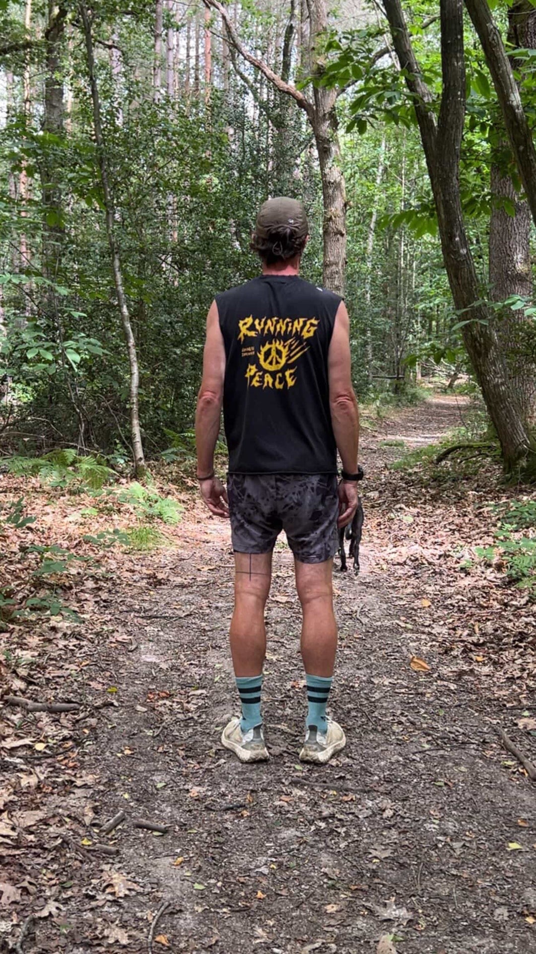 Man running on a trail in a forest wearing a black Nowhere Somewhere Running Peace tank top with yellow text.