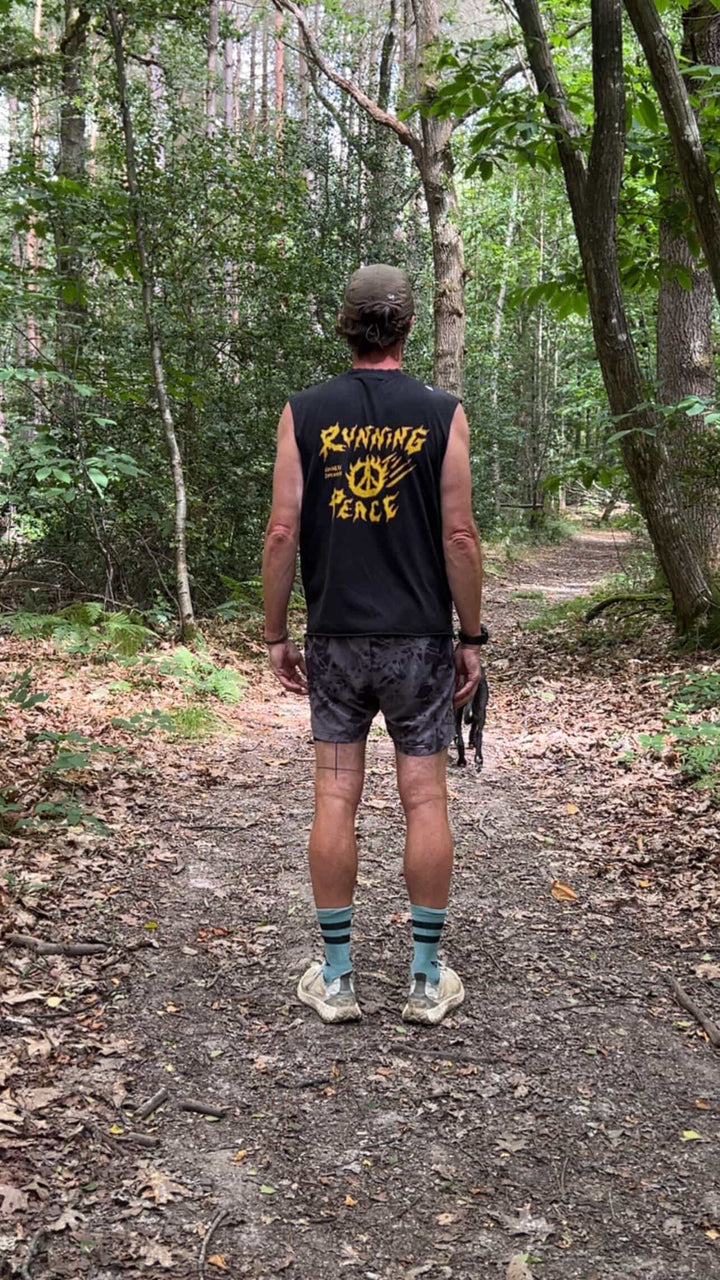 Man running on a trail in a forest wearing a black Nowhere Somewhere Running Peace tank top with yellow text.