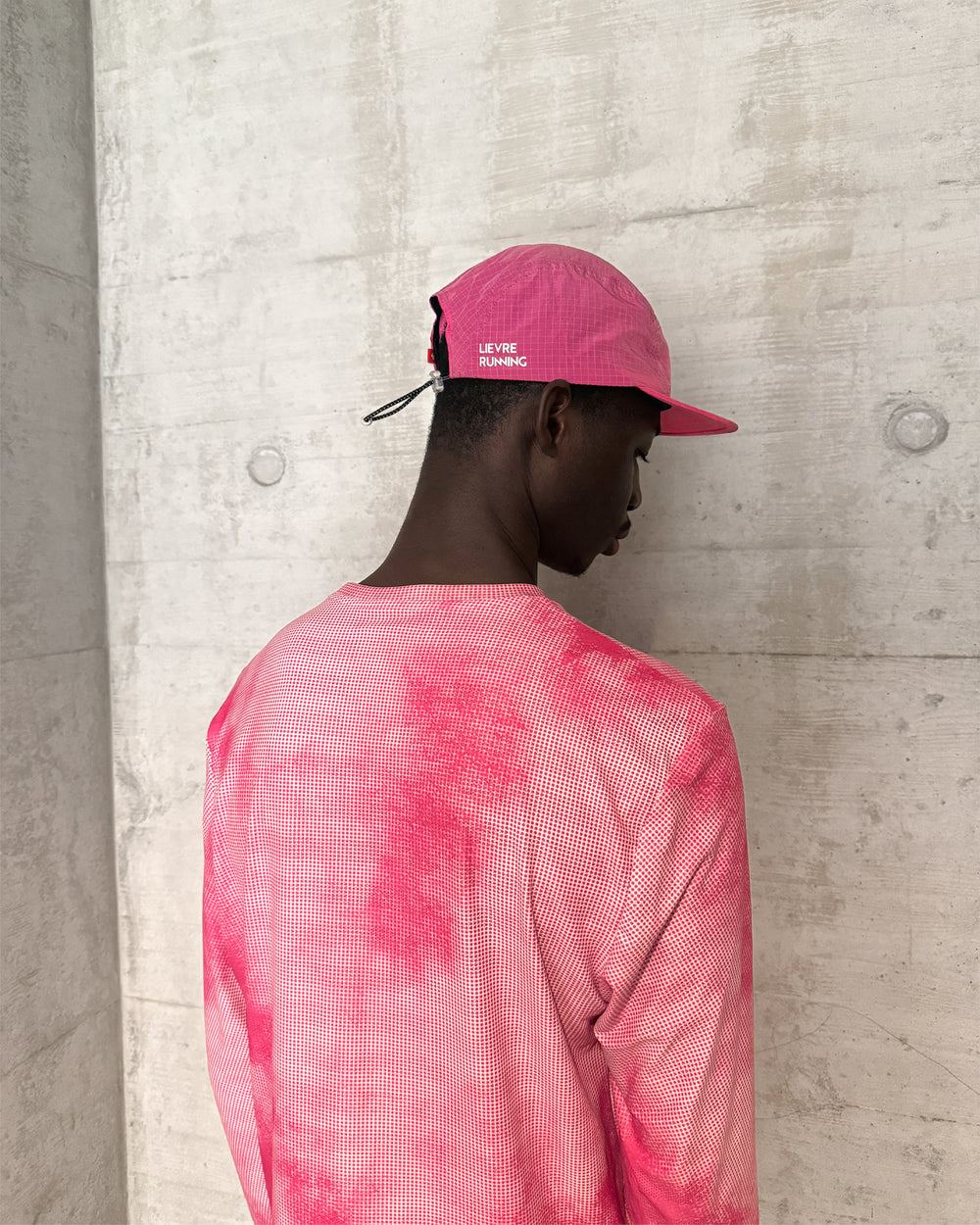 Person wearing a pink Lievre Runnin cap and matching shirt against a concrete wall
